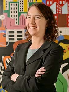 Professional portrait of a woman with crossed arms smiling, standing before a colorful mural of city buildings.