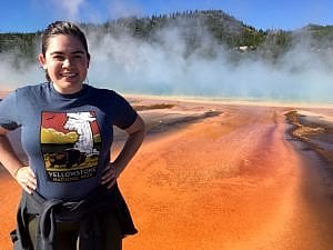Person standing in front of Grand Prismatic Spring in Yellowstone National Park, with colorful thermal features and steam rising in the background.
