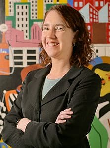 Professional portrait of a woman with crossed arms smiling, standing before a colorful mural of city buildings.