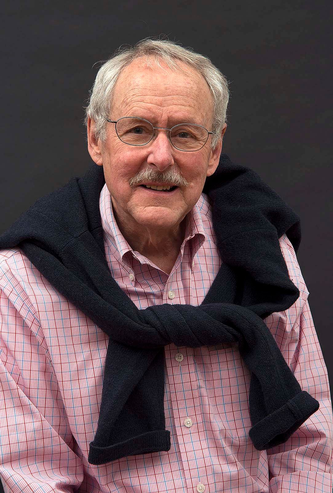 Portrait of Peter Hassrick with gray background. Pink and blue checked shirt with dark sweater tied over shoulders.