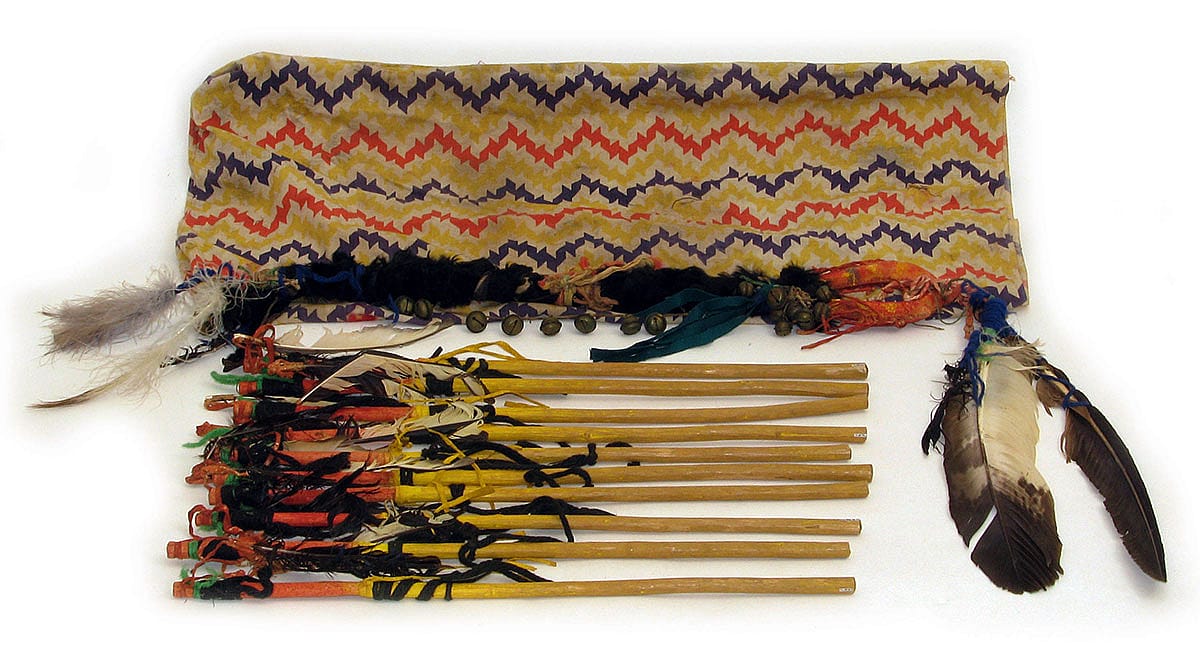 Hand game rattle with game sticks and carrying bag. Hide, feathers, leather, yarn, brass bells, wool cloth. The Paul Dyck Plains Indian Buffalo Culture Collection, acquired through the generosity of the Dyck family and additional gifts of the Nielson Family and the Estate of Margaret S. Coe. NA.503.36