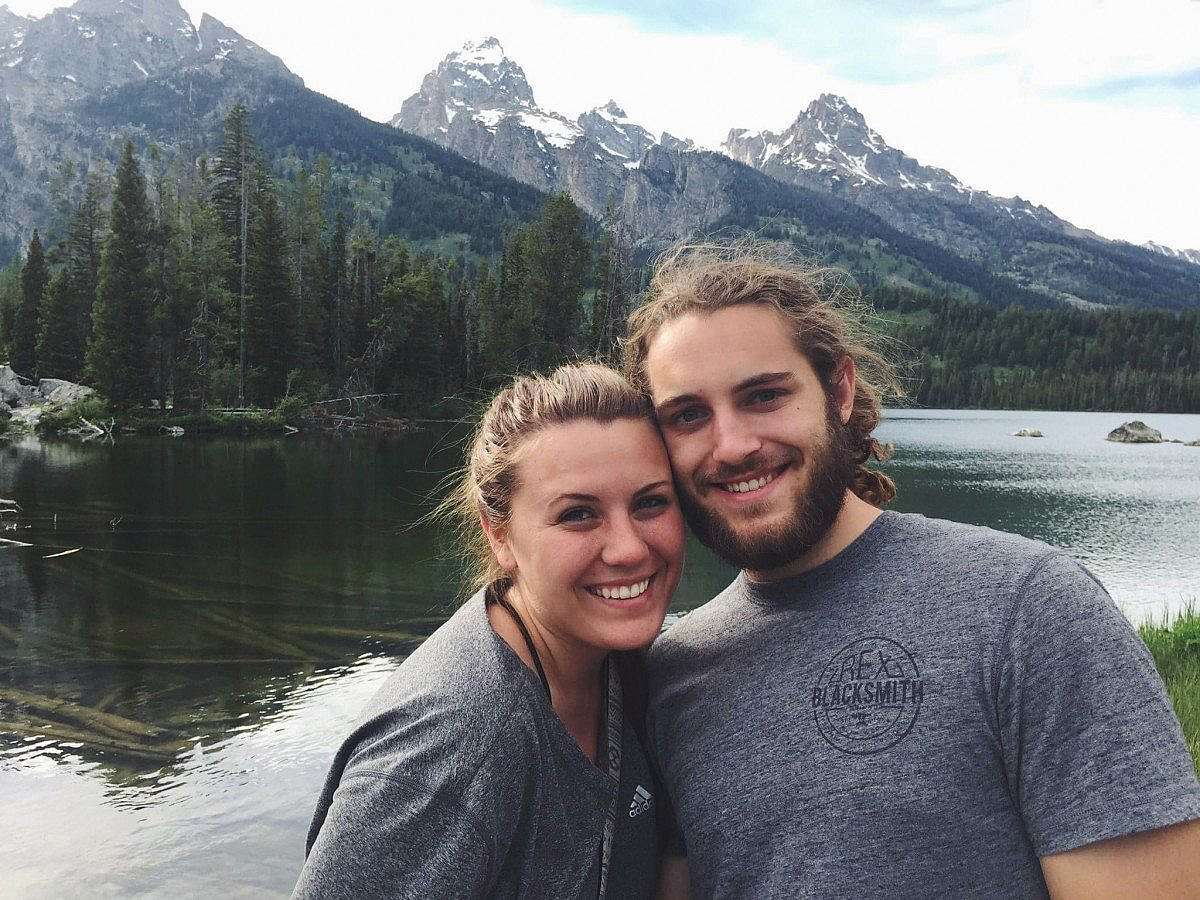 Smiling couple posing in front of Taggart Lake, with calm waters reflecting the towering, snow-capped peaks of the Grand Teton mountain range.
