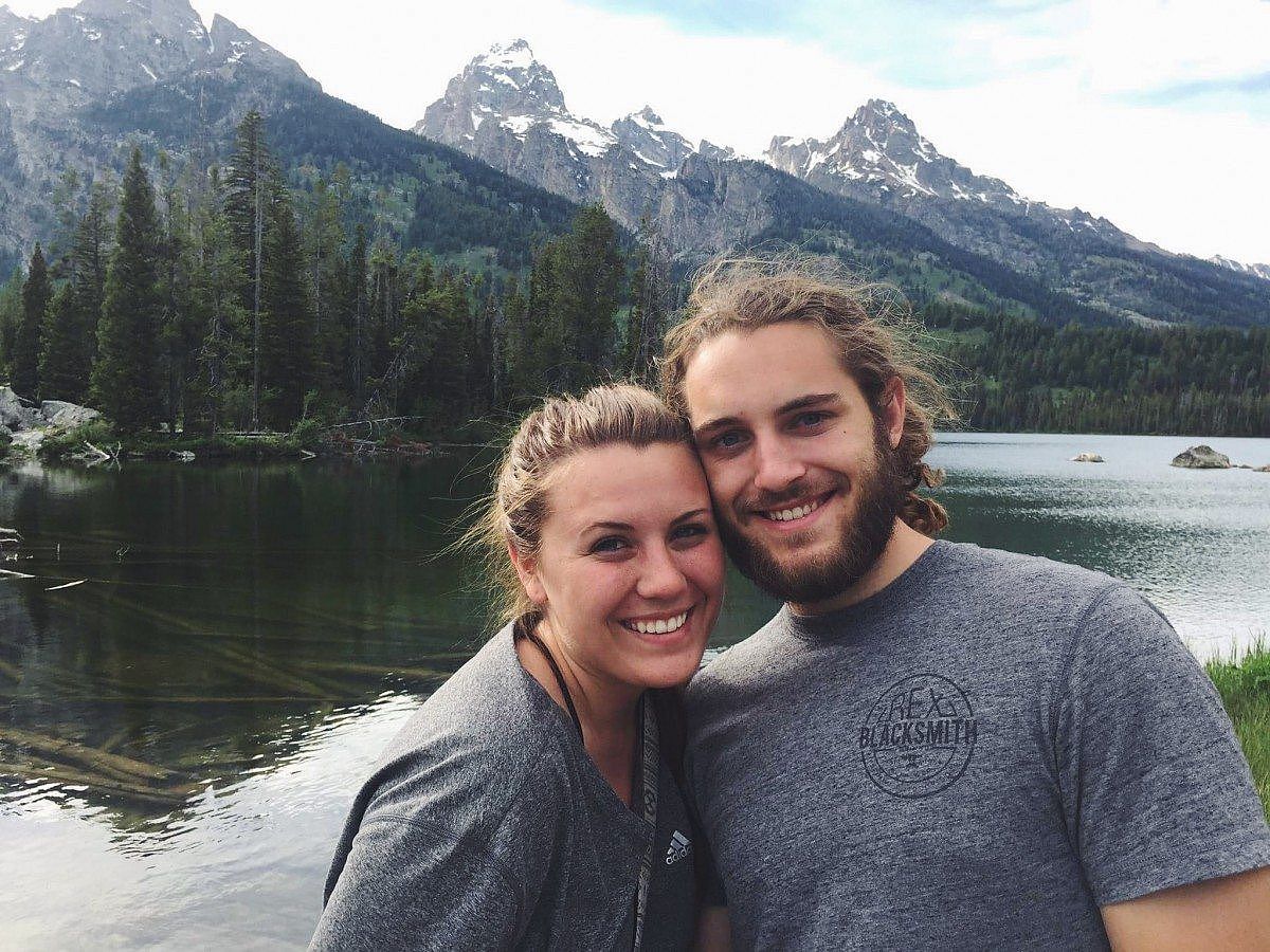 Smiling couple posing in front of Taggart Lake, with calm waters reflecting the towering, snow-capped peaks of the Grand Teton mountain range.