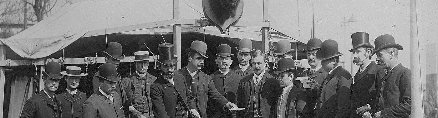 Black and white photograph. Group of men in hats and suits at Buffalo Bill's Wild West gather in front of William F. Cody's tent on Buffalo Bill's Wild West show backlot. Elliot & Fry, Baker Street, London, photographer. William F. Cody Collection. P.69.810 (detail)