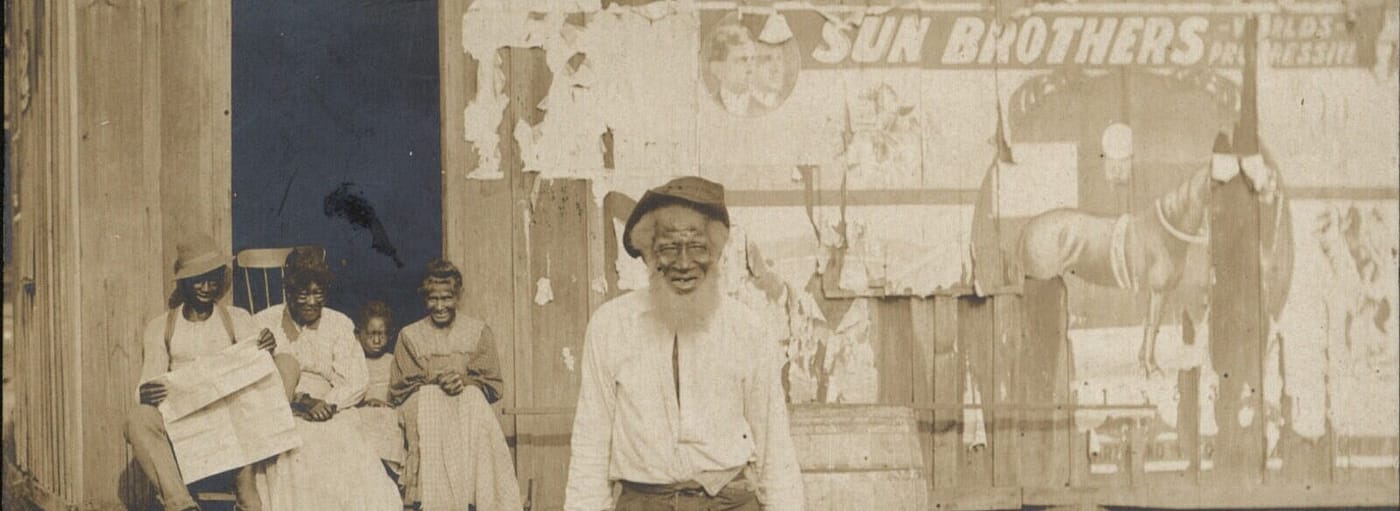 An unidentified elderly African American man stands in front of a building. A man, two women and a child sit on the porch of the building behind him
