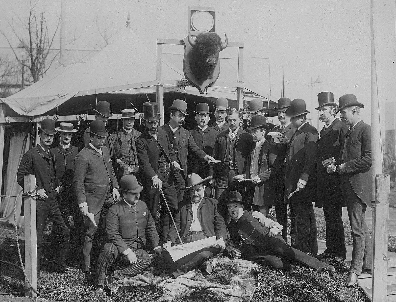 Black and white photograph. Group of businessmen in hats and suits gather in front of William F. Cody's tent on Buffalo Bill's Wild West show backlot. Elliot & Fry, Baker Street, London, photographer. William F. Cody Collection. P.69.810