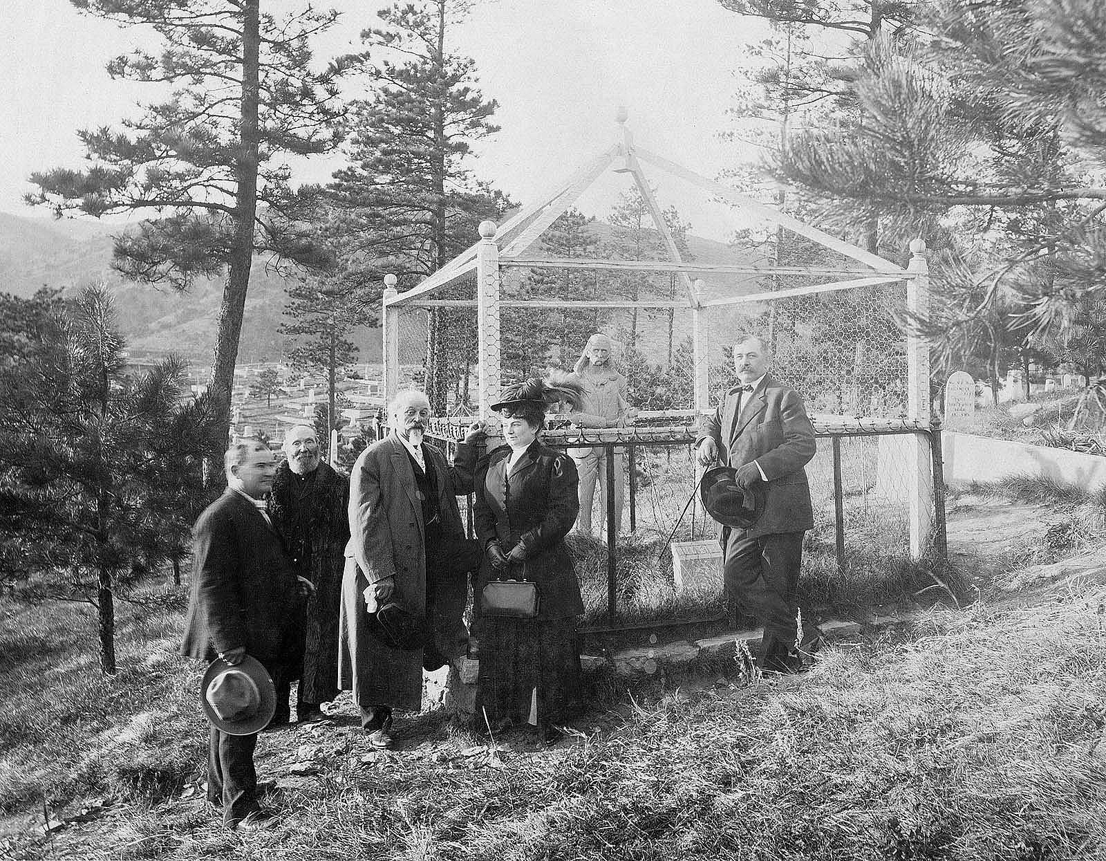 William F. "Buffalo Bill" Cody visits the grave of James Butler "Wild Bill" Hickok in Deadwood, South Dakota. A large statue of Hickok and a small marker are enclosed by a wrought iron fence and an enclosed wire fence.  Cemetery and mountains in background. MS 6 William F. Cody Collection. P.69.1069