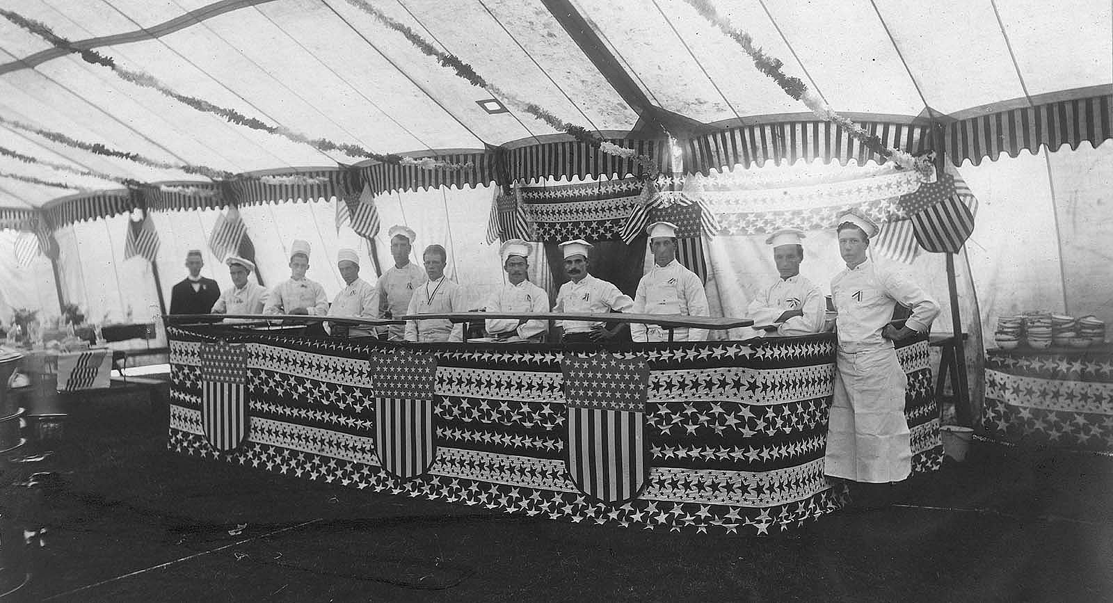 Several men in white clothes and chef's hats stand behind a bunting bedecked counter. Buffalo Bill's Wild West mess tent decorated for the Fourth of July, ca. 1890. P.69.0087
