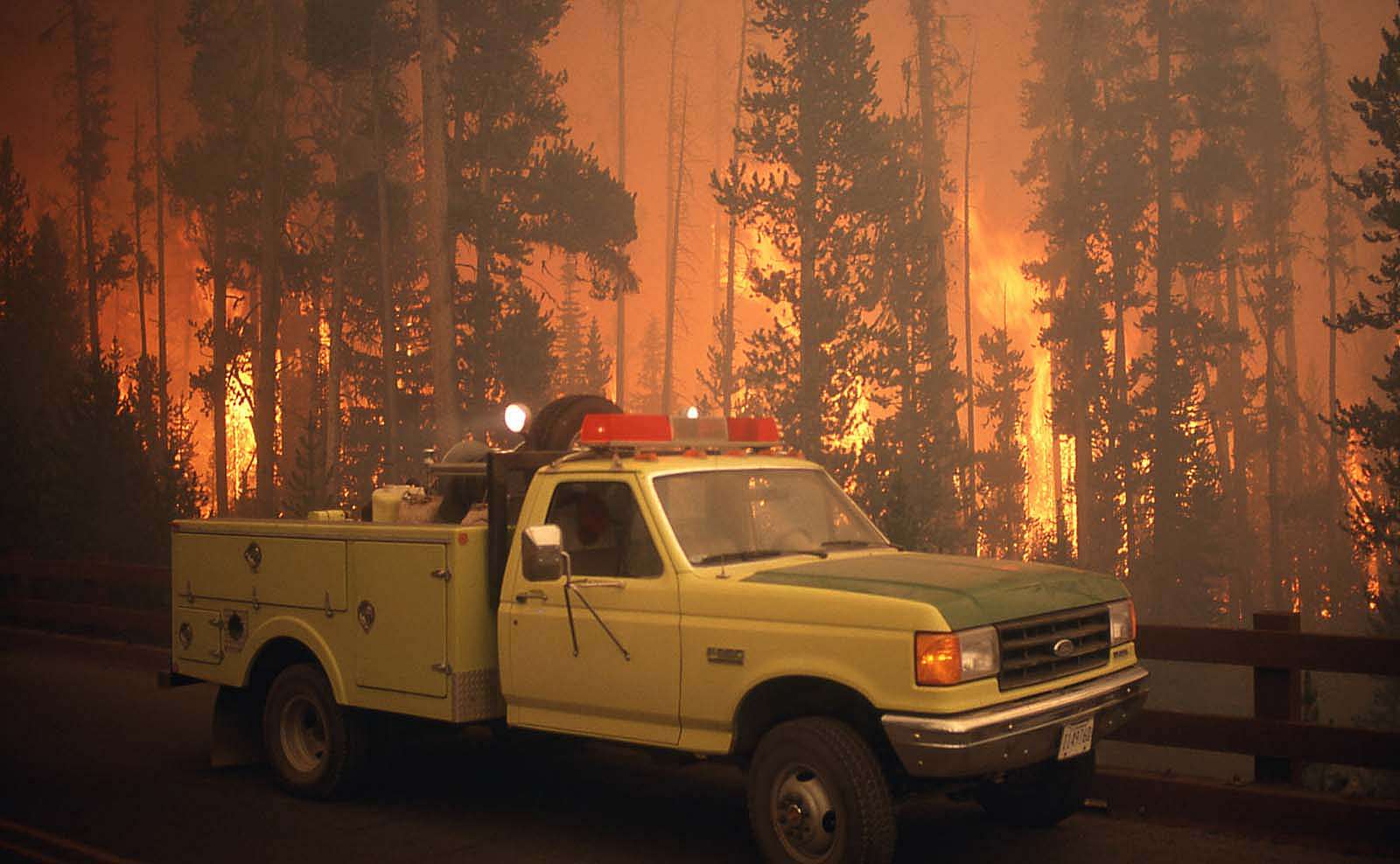 Yellow fire truck on bridge in the Grant Village campground with wildland fire in background. NPS photo by Jeff Henry, 1988.