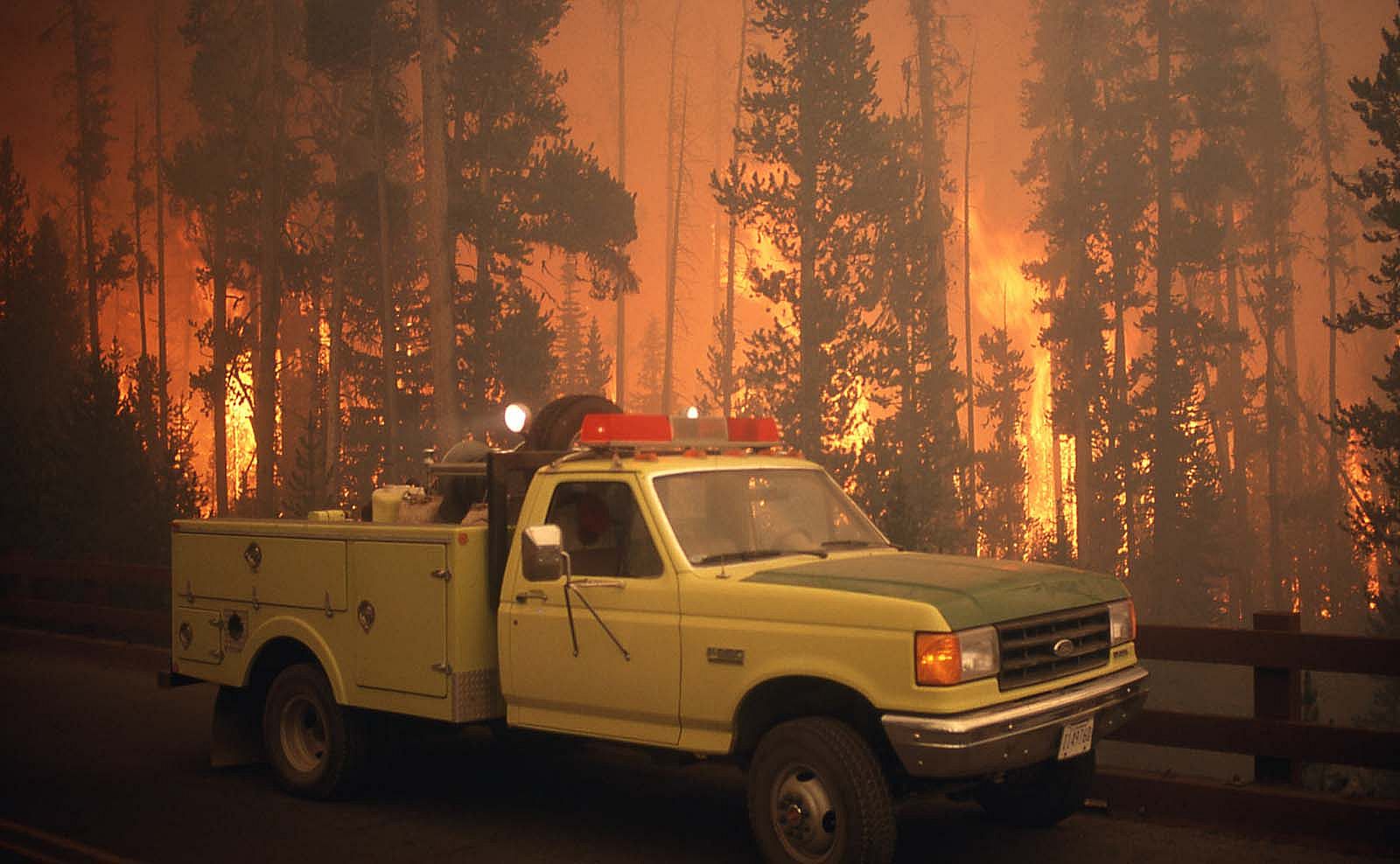 Yellow fire truck on bridge in the Grant Village campground with wildland fire in background. NPS photo by Jeff Henry, 1988.