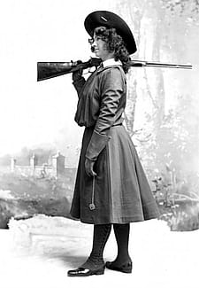 Woman in dress and wide-brimmed hat standing to side with rifle over shoulder.