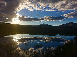 Scenic view of a sunset over a lake in Yellowstone National Park, with mountains and clouds reflecting on the water’s surface.