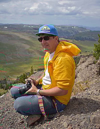 Brian Beauvais seated in high elevation landscape wearing blue ball cap and yellow windbreaker. He holds a camera. Photo by Corey Anco.