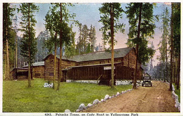 Photographic postcard featuring a colorized photograph of the Pahaska Tepee near Cody, Wyoming. The log structure is in the center. Trees are in the foreground and in the background. A car is parked in the drive on the right. MS 327 James Wojtowicz Collection, McCracken Research Library. MS327.08.03.008.01