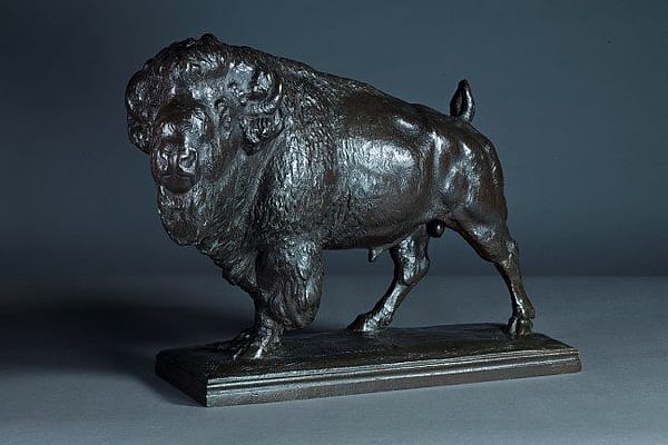 Bronze sculpture of a bison standing on a rectangular base, showcasing detailed musculature and textured fur against a dark background.