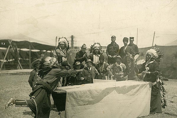 A performer who may be Oglala Lakota Chief Iron Tail plays ping pong with a fellow castmate as Joe Black Fox and a group Native performers, cowboys, vaqueros, and Cossacks watch the match. MS 6 William F. Cody Collection, McCracken Research Library. P.69.1031