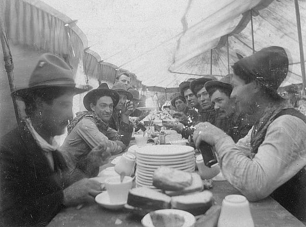 Group of cowboys from Buffalo Bill's Wild West sit at mess table with bread and stacks of plates on the table. MS 6 William F. Cody Collection. P.6.0213
