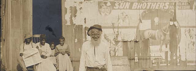 An unidentified elderly African American man stands in front of a building. A man, two women and a child sit on the porch of the building behind him