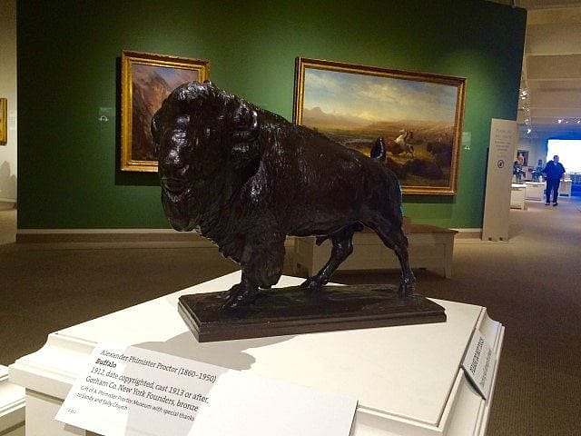 Bronze bison sculpture displayed in a museum, positioned on a pedestal with informational plaques, surrounded by framed paintings and exhibition lighting.