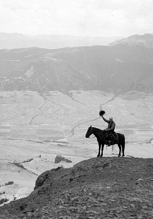 Ned Frost, astride his horse, waving his hat from the top of Jim Mountain, with the Frost & Richard Ranch in the Wapiti Valley behind and below him.