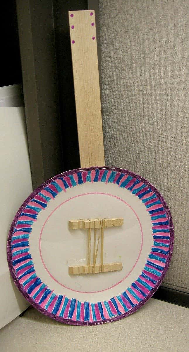 Handmade banjo crafted from a paper plate and colored yarn, featuring a simple wooden neck and strings.