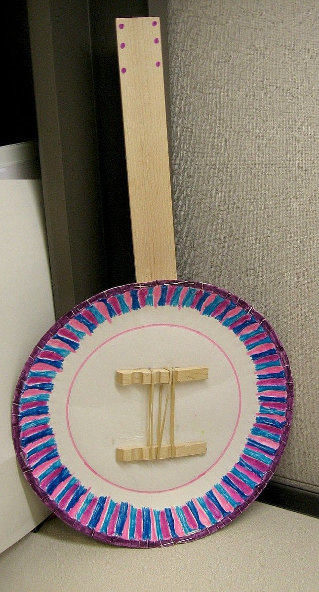 Handmade banjo crafted from a paper plate and colored yarn, featuring a simple wooden neck and strings.