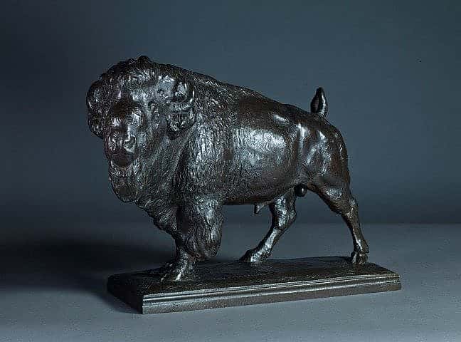 Bronze sculpture of a bison standing on a rectangular base, showcasing detailed musculature and textured fur against a dark background.