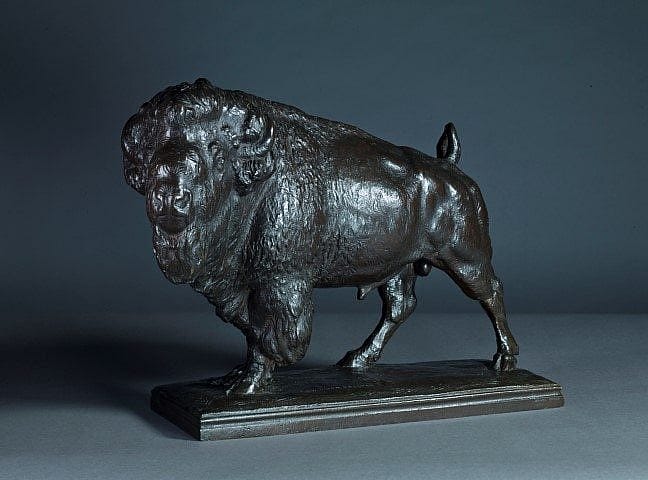 Bronze sculpture of a bison standing on a rectangular base, showcasing detailed musculature and textured fur against a dark background.