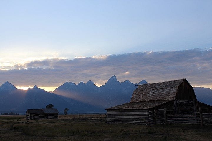 Historic wooden barns at Mormon Row silhouetted against the Grand Tetons, with golden light filtering through the clouds at sunset.