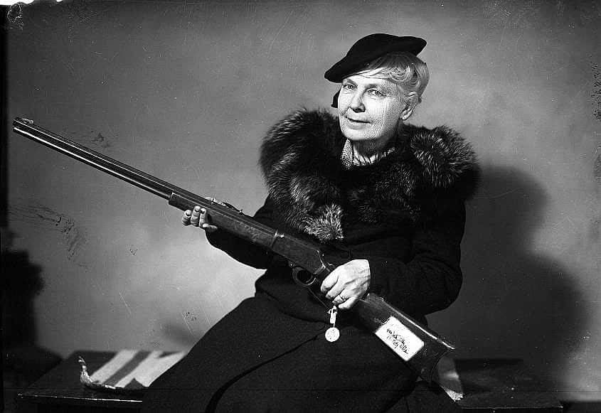 A black and white photo of an older lady wearing a fur coat and a hat, holding a long rifle.