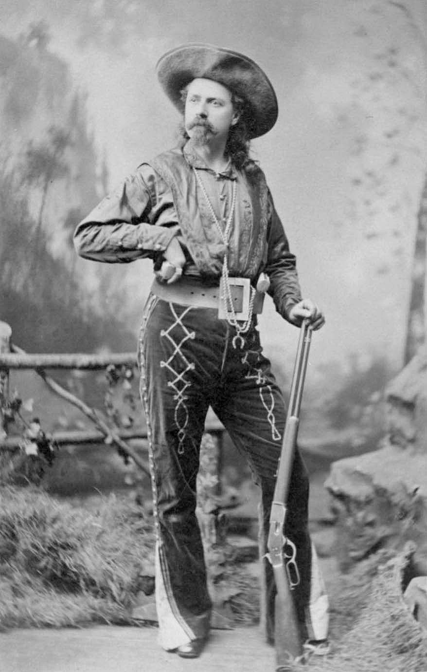 Man in cowboy show attire standing with rifle and pistols, wearing a cowboy hat.