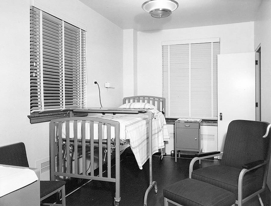 Black and white image of hospital with bed, chairs, and small bedside cabinet