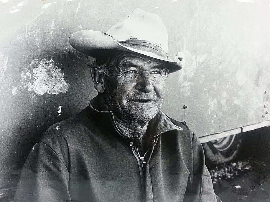 old man wearing cowboy hat leaning against trailer