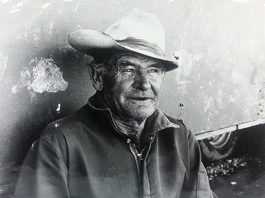 old man wearing cowboy hat leaning against trailer