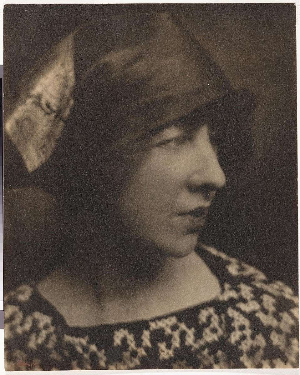 Vintage portrait of Gertrude Vanderbilt Whitney wearing a patterned dress and a stylish hat, in a sepia tone.