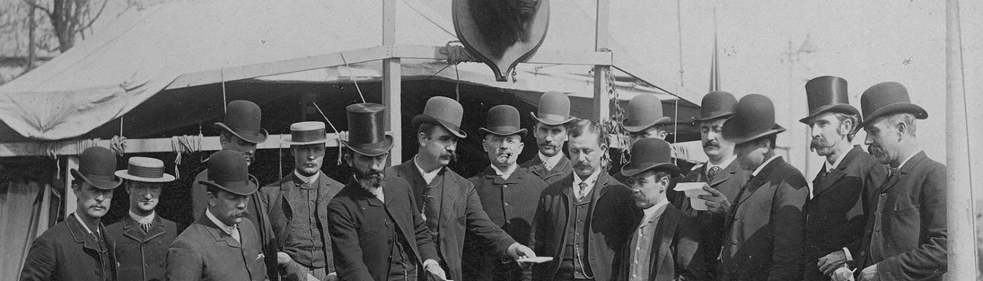 Black and white photograph. Group of men in hats and suits at Buffalo Bill's Wild West gather in front of William F. Cody's tent on Buffalo Bill's Wild West show backlot. Elliot & Fry, Baker Street, London, photographer. William F. Cody Collection. P.69.810 (detail)