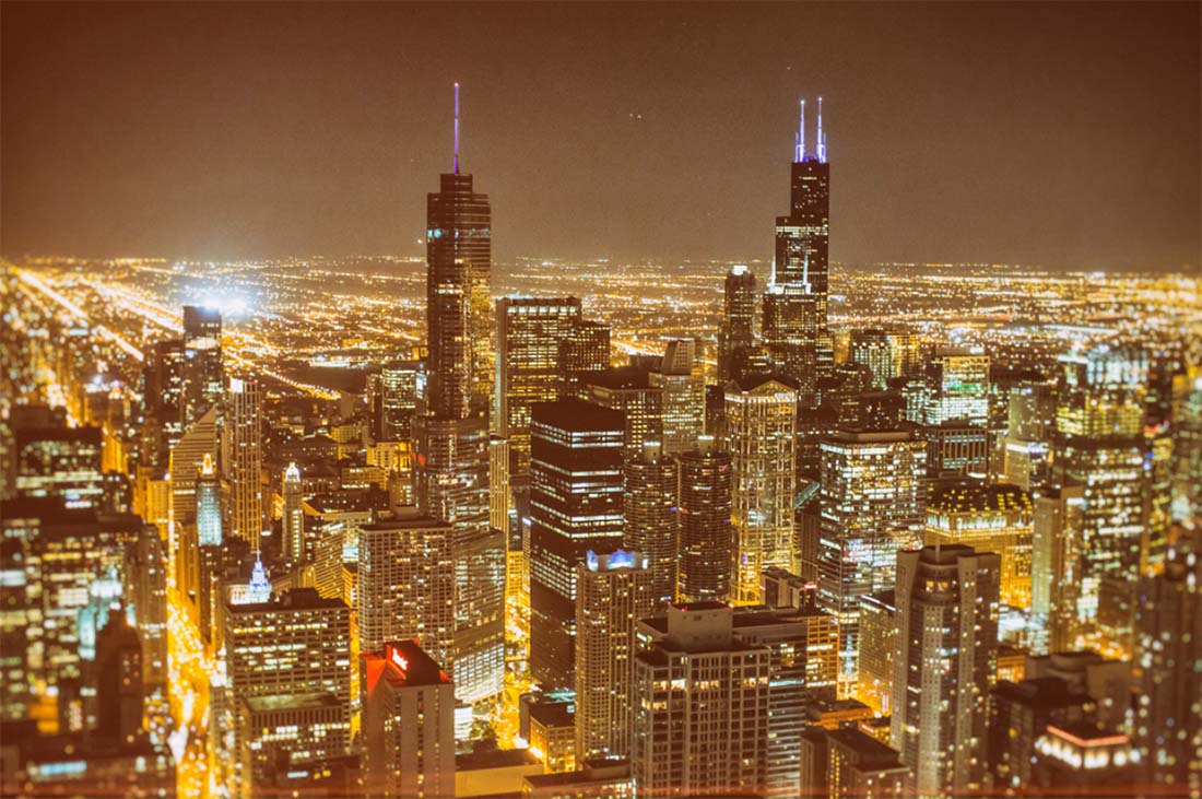 A night scene with bright lights of tall buildings in the foreground and other lit buildings fading off into the distance, giving the entire photo a yellow glow.